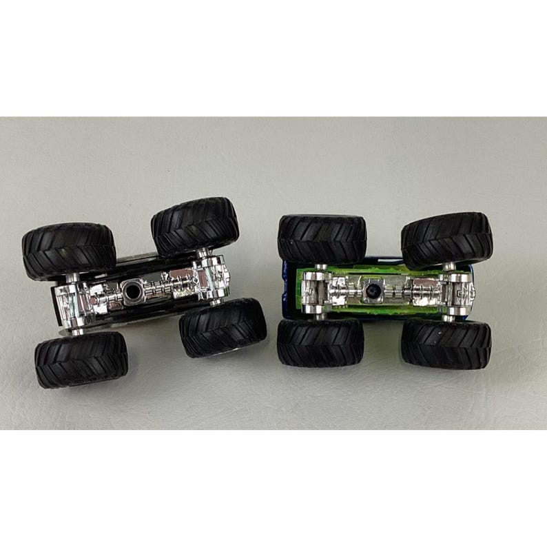 K&ouml;nnte beinhalten: Zwei Spielzeug-Monstertrucks, einer schwarz und einer blau, sind von unten zu sehen. Jeder Truck hat gro&szlig;e, schwarze, grob profilierte Reifen und ein detailliertes silbernes Fahrgestell. Die Trucks stehen auf einer hellgrauen Oberfl&auml;che.