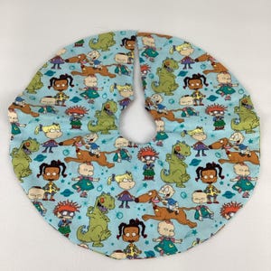May include: A light blue, circular tree skirt with a repeating pattern of cartoon characters and dinosaurs. The skirt has a split opening for the tree trunk and is made of fabric. The design includes characters in various outfits and poses.