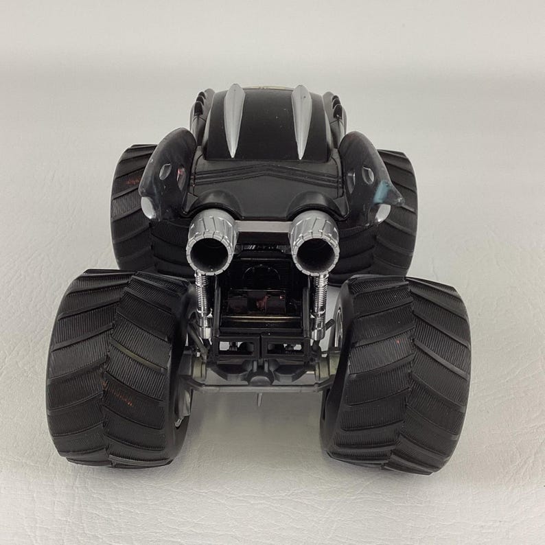May include: A black monster truck toy with large, textured tires. The truck has silver exhaust pipes and a silver accent on the top. The toy is designed with a sleek, aerodynamic body.