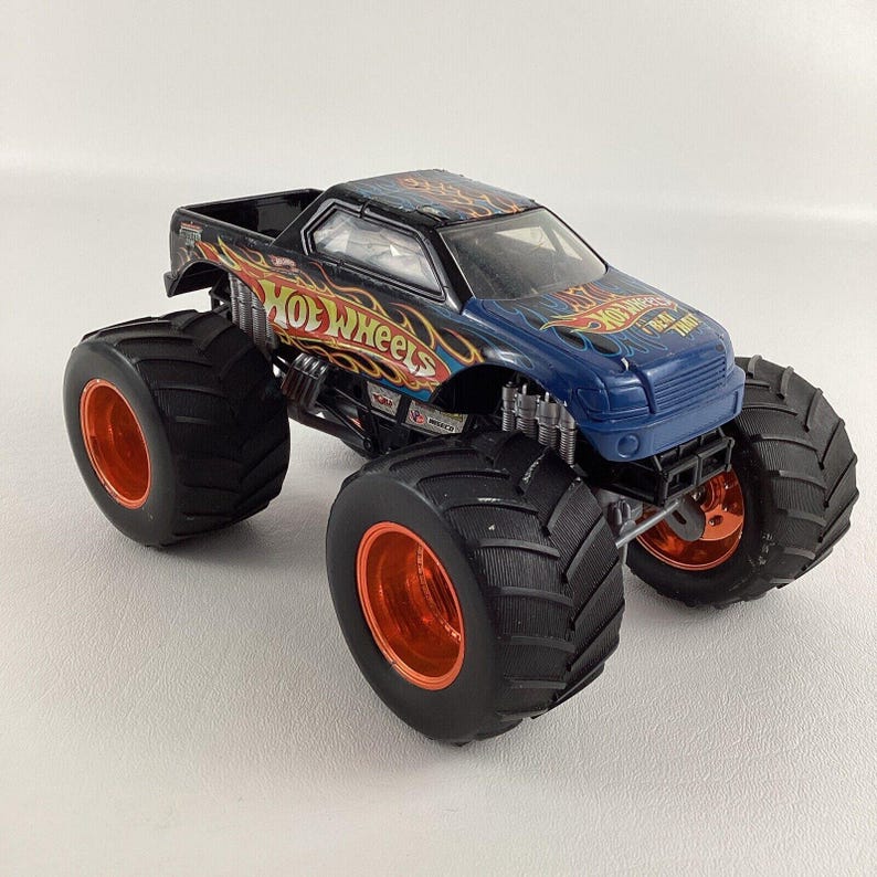 Puede incluir: Un juguete de cami&oacute;n monstruo Hot Wheels en negro y azul con detalles en naranja. El cami&oacute;n presenta neum&aacute;ticos negros grandes con llantas naranjas, y el logotipo de Hot Wheels es visible. El cami&oacute;n tiene un dise&ntilde;o detallado con llamas.