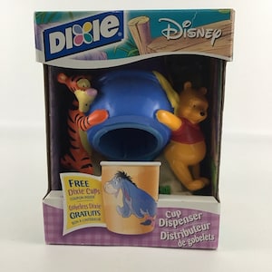 May include: A Winnie the Pooh themed cup dispenser with a blue base and a Winnie the Pooh character on the front. The dispenser is made of plastic and has a clear plastic cup holder. The box has a blue and white checkered pattern and the text "FREE Dixie Cups COUPON INSIDE Gobelets Dixie GRATUITS BON À L'INTÉRIEUR" on the front.