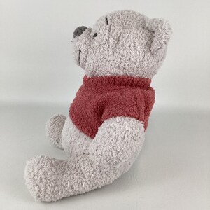 May include: A plush teddy bear in a seated position, featuring a light gray body and a red sweater. The bear has a gray nose and is made of a soft, textured material, perfect for cuddling. A classic children's toy.