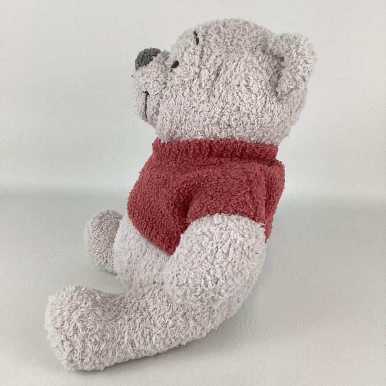 May include: A plush teddy bear in a seated position, featuring a light gray body and a red sweater. The bear has a gray nose and is made of a soft, textured material, perfect for cuddling. A classic children's toy.