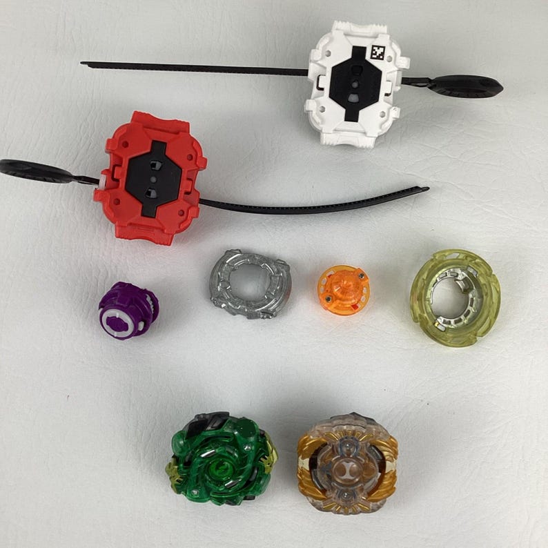 May include: An assortment of colorful spinning top toys and launchers. The image shows a red and white launcher with a black pull string, along with various tops in green, gold, purple, silver, and orange. The toys are designed for competitive play.