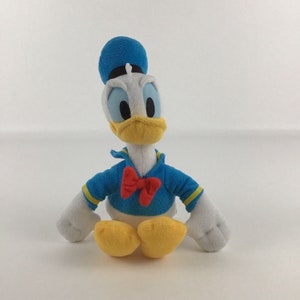 May include: A plush toy of Donald Duck wearing a blue sailor hat and a red bow tie. He is sitting with his legs crossed and has a yellow beak and white feathers.