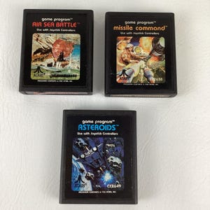 May include: Three black rectangular Atari game cartridges featuring labels for Air Sea Battle, Missile Command, and Asteroids. Each label includes the game title, the text game program, Use with Joystick Controllers, and the Atari logo.