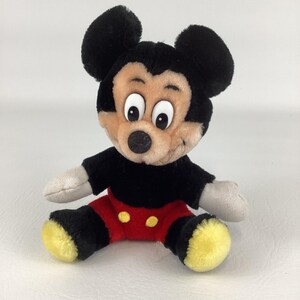 May include: A plush Mickey Mouse toy, featuring a black body, red shorts with yellow buttons, and large black ears. The toy has a tan face with big white eyes and a black nose. The arms and hands are gray. The toy is sitting.