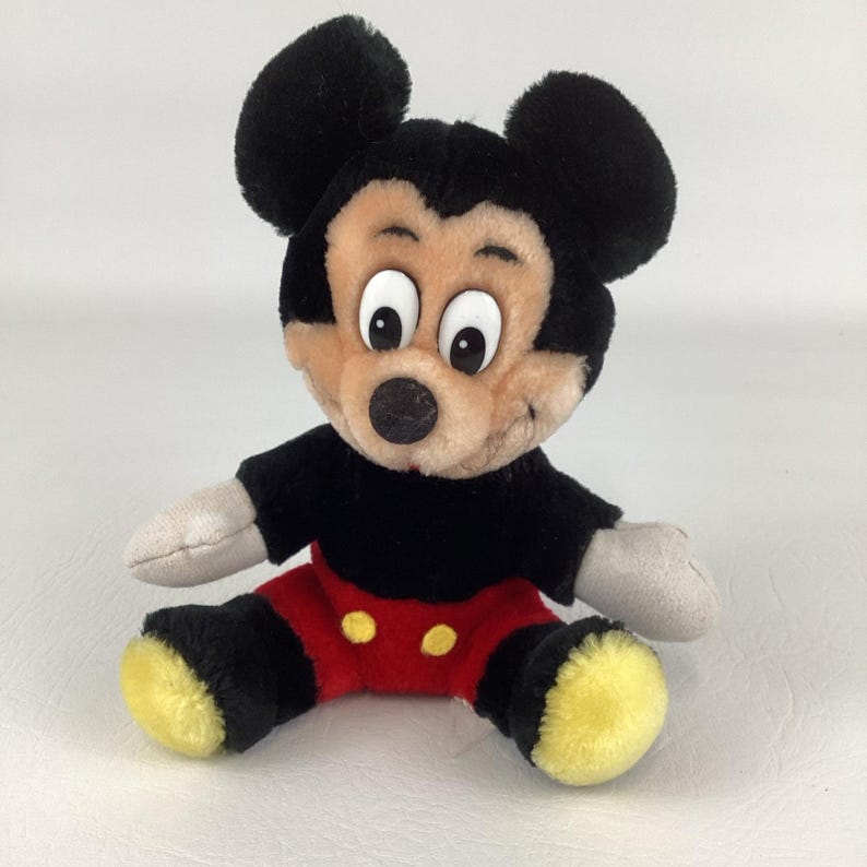 May include: A plush Mickey Mouse toy, featuring a black body, red shorts with yellow buttons, and large black ears. The toy has a tan face with big white eyes and a black nose. The arms and hands are gray. The toy is sitting.