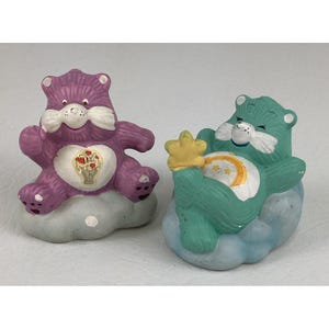 May include: Two Care Bear figurines. One is purple with a heart design, the other is teal with a rainbow and star design. Both are sitting on cloud-shaped bases. The figurines are vintage and have a painted finish.