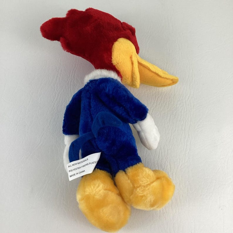 Op de afbeelding: Een pluche Woody Woodpecker pop, met een rode kop, gele snavel en blauw lichaam. De pop heeft witte handschoenen en gele voeten. Een label luidt "ALL NEW MATERIALS POLYESTER FIBERS PLASTIC MADE IN CHINA".