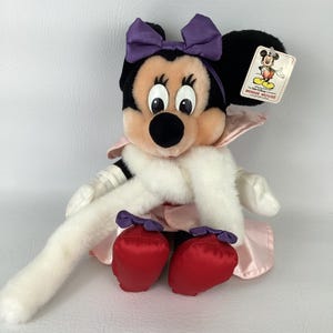 May include: A plush Minnie Mouse doll featuring a purple satin bow, a long white faux fur stole, a light pink satin coat, and red satin shoes. The doll has black ears, a peach-coloured face, and a white tag attached to the ear reading MINNIE MOUSE.