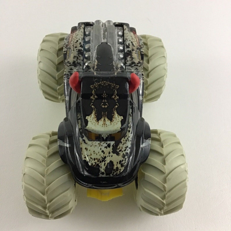 May include: A black monster truck toy with a camouflage pattern and red horns. The truck has large, white tires and a detailed engine.