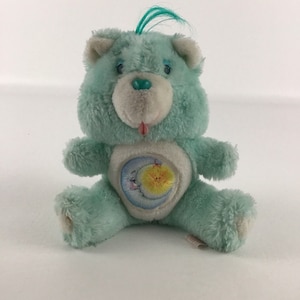 Peut inclure: Un ours en peluche bleu clair Care Bear avec une lune et une étoile sur le ventre. L'ours a un visage blanc avec un nez bleu et des yeux bleus. L'ours a une langue bleue qui sort.