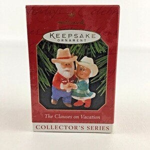 May include: A red box with a white border and the words "Keepsake Ornament" in white text. The box features a picture of a Santa Claus and Mrs. Claus dressed in cowboy attire. The text "The Clauses on Vacation" is printed below the image. The words "Collector's Series" are printed in white text on a red background at the bottom of the box.