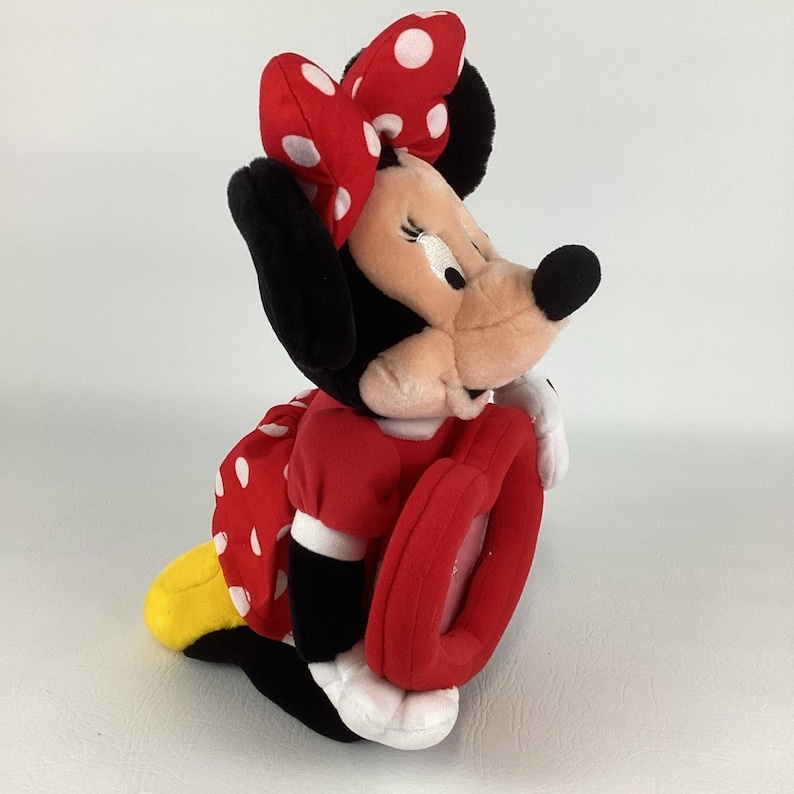 Pu&ograve; includere: Bambola di peluche di Minnie Mouse seduta. Indossa un vestito rosso a pois bianchi, un fiocco abbinato e guanti bianchi. Le sue orecchie e il naso neri sono prominenti e si vedono le sue scarpe gialle.