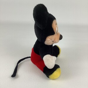 May include: A plush Mickey Mouse toy, featuring a black body, ears, and tail, with red shorts, white gloves, and yellow shoes. The iconic character is seated, showcasing its classic design and colors.