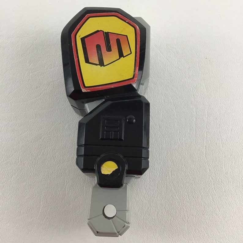 May include: Black and gray plastic toy with a yellow and red logo that says "SM" on it. The toy has a gray handle and a black body.