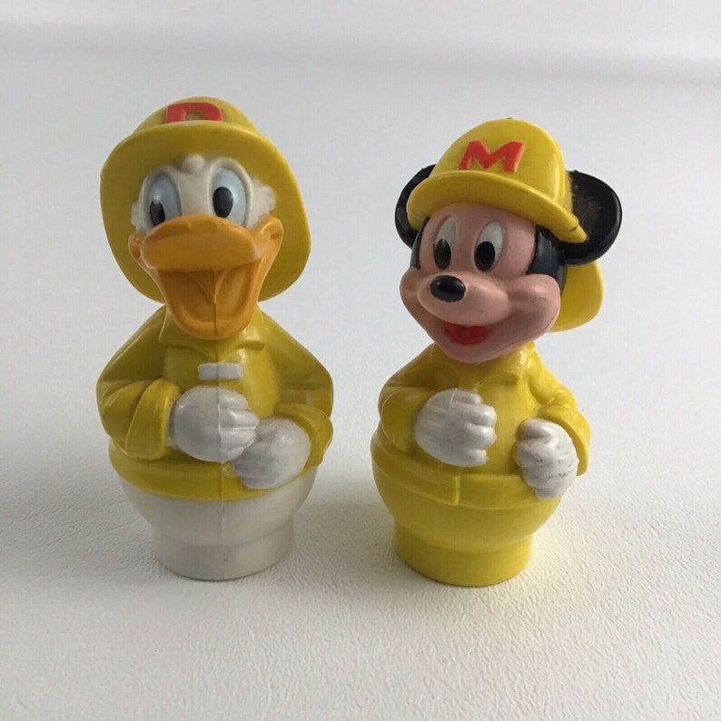 May include: Two yellow plastic figurines of Mickey Mouse and Donald Duck dressed as firefighters. Mickey Mouse is wearing a yellow raincoat and a red hat with a white "M" on it. Donald Duck is wearing a yellow raincoat and a red hat with a white "D" on it.