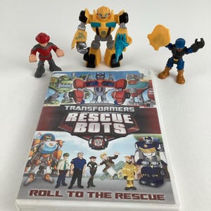 Op de afbeelding: Een DVD met de animatieserie Transformers Rescue Bots. De DVD-hoes heeft een cartoonafbeelding van de Rescue Bots-personages, waaronder Optimus Prime, Heatwave, Blades, Chase en Boulder. De titel van de DVD is "Roll to the Rescue". De afbeelding bevat ook drie kleine actionfiguren: een gele Bumblebee, een rode brandweerman en een blauwe superheld.