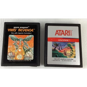 May include: Two black rectangular Atari 2600 video game cartridges. The left cartridge features the Yars' Revenge game title, a winged insect illustration, and the Atari logo. The right cartridge displays the Centipede game title and artwork.