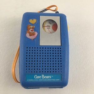 May include: A blue Care Bears portable radio with an orange strap. The radio features a Care Bear sticker on the front and a tuning dial. The bottom of the radio has the Care Bears logo and the American Greetings Corp. copyright information.