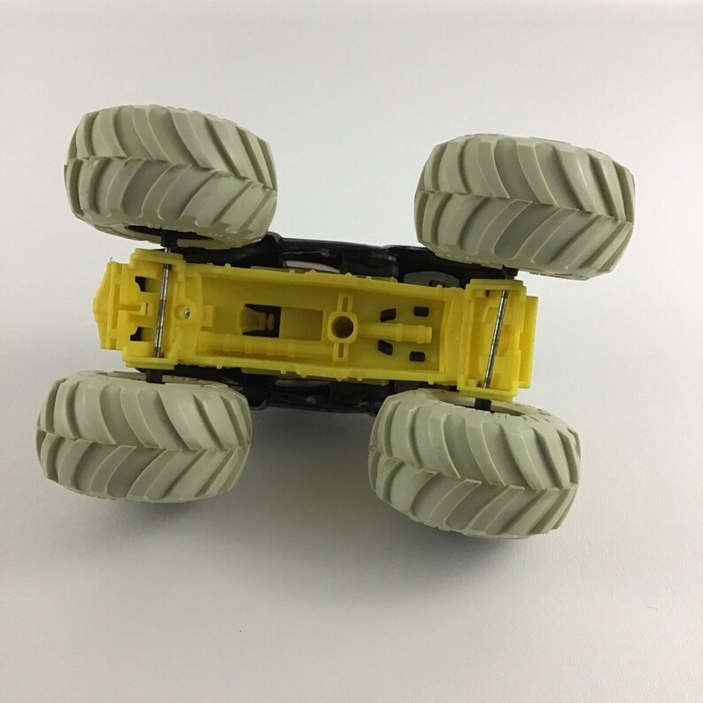 May include: A yellow toy monster truck with four large gray wheels. The truck is viewed from the bottom, showing the underside of the chassis.