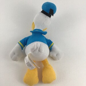 May include: A white plush toy of Donald Duck wearing a blue sailor shirt with yellow trim, a blue sailor hat with a black band, and yellow feet.