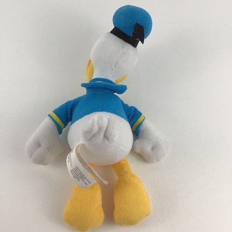 May include: A white plush toy of Donald Duck wearing a blue sailor shirt with yellow trim, a blue sailor hat with a black band, and yellow feet.