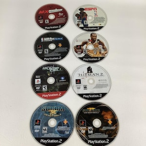 May include: Eight circular PlayStation 2 game discs arranged in two columns. Titles include MX Rider, ESPN NBA 2K5, NBA 08, NBA Live 08, Tom Clancy's Splinter Cell, Hitman 2, SOCOM U.S. Navy SEALs, and SOCOM 3 U.S. Navy SEALs, featuring various graphics.