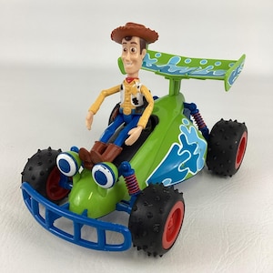 May include: A green and blue toy race car with a Woody figure from Toy Story. The car has large black tires with red rims, blue accents, and a green spoiler. Woody is wearing a cowboy hat, yellow shirt, blue pants, and brown boots.