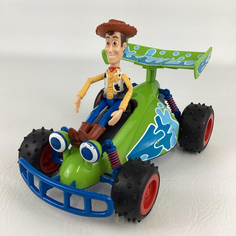 May include: A green and blue toy race car with a Woody figure from Toy Story. The car has large black tires with red rims, blue accents, and a green spoiler. Woody is wearing a cowboy hat, yellow shirt, blue pants, and brown boots.