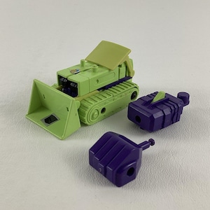 May include: A green toy bulldozer with a purple arm and claw. The bulldozer has a black track and a yellow sticker on the side. The arm and claw are separate pieces.