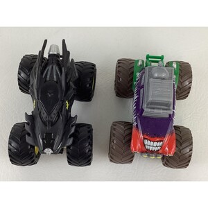 May include: Two toy monster trucks. One is black with yellow accents, resembling Batman's vehicle. The other is purple, red, and green, with a grinning face design, reminiscent of the Joker.