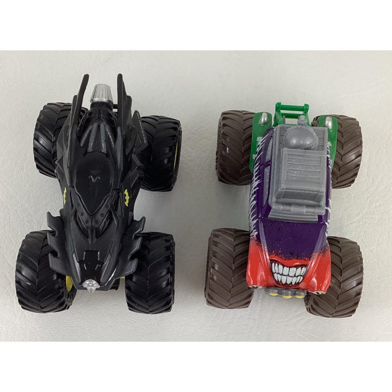 May include: Two toy monster trucks. One is black with yellow accents, resembling Batman's vehicle. The other is purple, red, and green, with a grinning face design, reminiscent of the Joker.