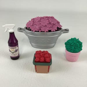May include: A collection of miniature gardening items, including a silver flower pot filled with pink flowers, a small green plant in a pink pot, a pot of red berries, and a purple spray bottle labeled "Linen Spray".