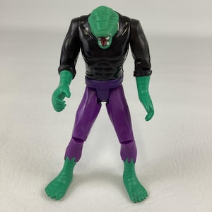 Puede incluir: Una figura de acci&oacute;n de pl&aacute;stico con cabeza de reptil verde y lengua roja, camiseta negra de manga larga con m&uacute;sculos abdominales moldeados y pantalones morados. Las manos y los pies verdes tienen detalles texturizados tipo vendaje.