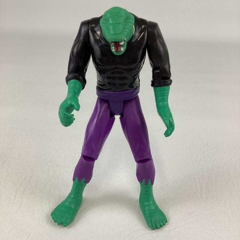 Puede incluir: Una figura de acci&oacute;n de pl&aacute;stico con cabeza de reptil verde y lengua roja, camiseta negra de manga larga con m&uacute;sculos abdominales moldeados y pantalones morados. Las manos y los pies verdes tienen detalles texturizados tipo vendaje.