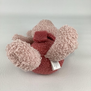 May include: A plush toy in shades of pink and red. The toy has a soft, fuzzy texture and a rounded shape. The toy's ears and paws are a lighter pink, while the body is a darker red. A tag is attached to the toy.