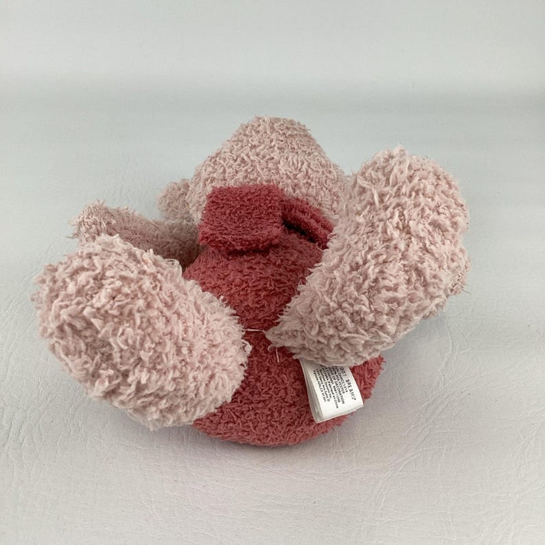May include: A plush toy in shades of pink and red. The toy has a soft, fuzzy texture and a rounded shape. The toy's ears and paws are a lighter pink, while the body is a darker red. A tag is attached to the toy.