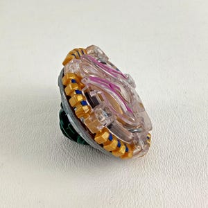 Puede incluir: Un juguete de peonza transparente y dorado con una parte superior transparente y un borde dorado y azul. El centro presenta detalles en rosa. La base es verde y plateada.