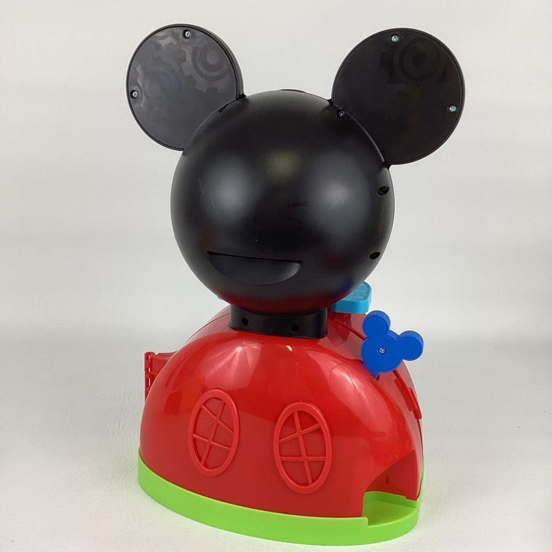 Pu&ograve; includere: Un giocattolo rosso e nero a tema Mickey Mouse con testa e orecchie nere, corpo rosso con due finestre ovali e base verde. Il giocattolo ha un pulsante blu a forma di Mickey Mouse, probabilmente un giocattolo per bambini.
