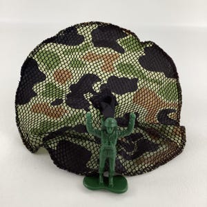 May include: A green plastic toy soldier stands beneath a camouflage net. The net features a pattern of black, brown, and green spots. The toy soldier is in a standing position with arms raised.