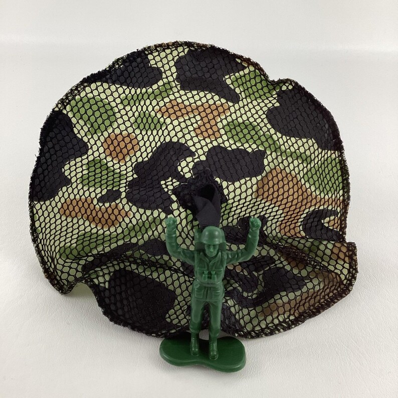 May include: A green plastic toy soldier stands beneath a camouflage net. The net features a pattern of black, brown, and green spots. The toy soldier is in a standing position with arms raised.