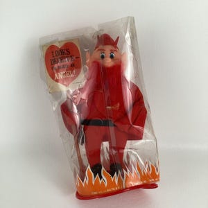 May include: A red devil doll with a black belt and horns, holding a stick. The doll is packaged in plastic with a red heart-shaped sign that reads "Looks Deceptive - I'm really an Angel!"