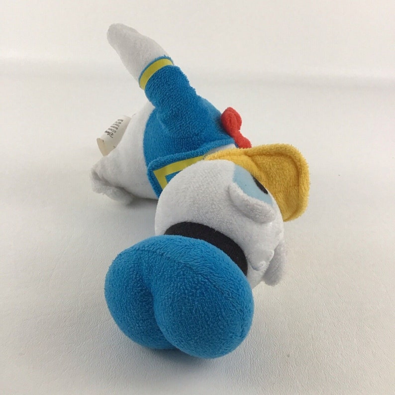 May include: A white plush toy of Donald Duck lying on its side. The toy is wearing a blue sailor shirt with yellow trim, a yellow hat, and a red bow tie. The toy has a blue hat and a white face with black eyes.
