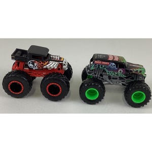 May include: Two toy monster trucks. One is red and black with a skull graphic and flames. The other is green and black with a skull graphic and the words "Grave Digger". Both have large black tires.