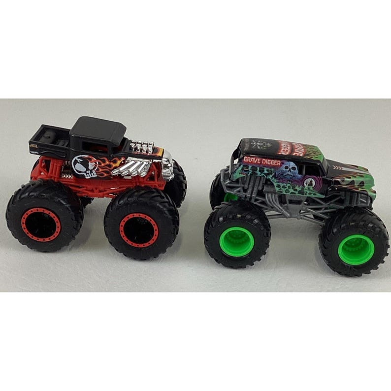 May include: Two toy monster trucks. One is red and black with a skull graphic and flames. The other is green and black with a skull graphic and the words "Grave Digger". Both have large black tires.