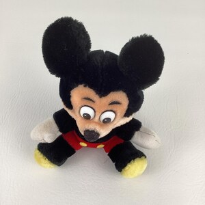 May include: A plush Mickey Mouse toy with large black ears, a black body, red shorts, and yellow feet. The toy has a tan face with large, wide eyes and a black nose. The toy is lying on a white surface.