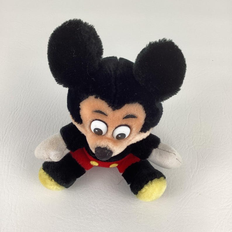May include: A plush Mickey Mouse toy with large black ears, a black body, red shorts, and yellow feet. The toy has a tan face with large, wide eyes and a black nose. The toy is lying on a white surface.