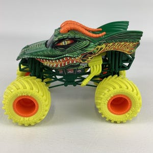 Pu&ograve; includere: Un camion mostro giocattolo verde e arancione con un design a drago. Il camion ha grandi pneumatici gialli con centri arancioni e una testa di drago dettagliata. &Egrave; un veicolo giocattolo.